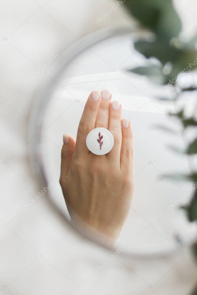 Stylish modern white round ring on beautiful hand and eucalyptus branch reflected in mirror on soft white tulle. Unusual fashionable fused glass ring on female hand with white manicure
