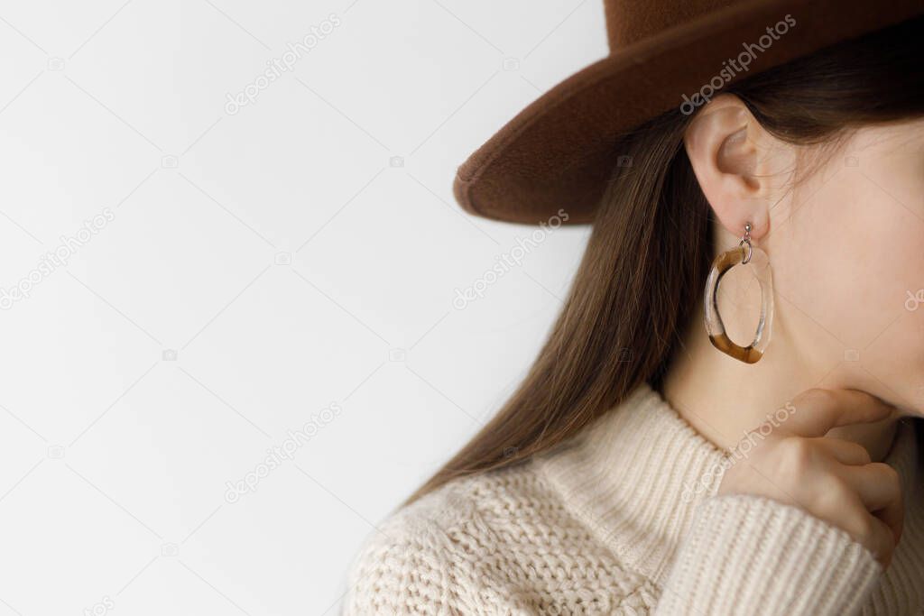 Beautiful stylish woman in hat with modern boho earring, cropped view. Fashionable female in sweater with unusual fused glass accessory. Beauty and care concept. Space for text