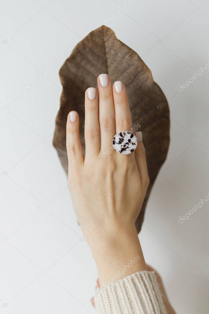 Stylish modern round ring on beautiful hand on background of brown dried big leaf on white wall. Unusual fashionable fused glass ring on female hand with white manicure. Care concept