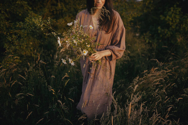 Beautiful woman in linen dress gathering wildflowers in summer meadow in sunny evening. Stylish young female in rustic dress picking flowers in countryside. Slow life. Atmospheric rural moment