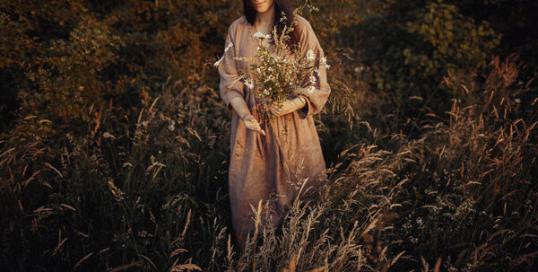 Beautiful woman in linen dress gathering wildflowers in summer meadow in evening. Stylish young female in rustic dress picking flowers in countryside. Atmospheric stylish vintage image