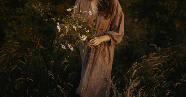Beautiful woman in linen dress gathering wildflowers in summer meadow in evening. Atmospheric stylish vintage image. Stylish young female in rustic dress picking flowers in countryside
