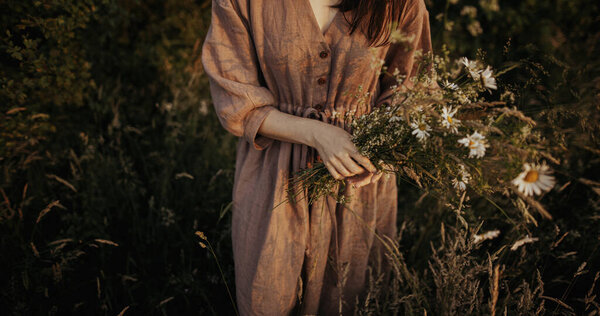 Beautiful woman in linen dress gathering wildflowers in summer meadow in evening. Stylish young female in rustic dress picking flowers in countryside. Atmospheric stylish vintage image