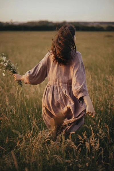 美丽的女人穿着亚麻布衣服 手牵着野花在夏夜的草地上奔跑 在乡村 穿着乡村风情服装的时髦年轻女性享受着免费的夜晚 无忧无虑的时刻 — 图库照片