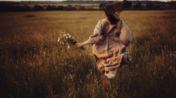 Beautiful woman in linen dress running with wildflowers in hand in summer meadow in sunset. Stylish young female in rustic dress enjoying freedom in countryside. Atmospheric stylish vintage image