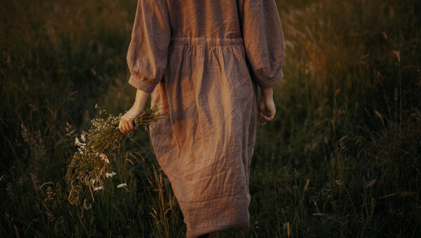Beautiful woman in linen dress walking with wildflowers in hand in summer meadow in sunset. Atmospheric stylish vintage image. Young female in rustic dress relaxing in evening in countryside