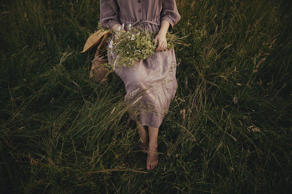 Beautiful woman in linen dress sitting barefoot on rustic chair among grass with bouquet of wildflowers in summer meadow. Young female relaxing in countryside. Atmospheric moment. Top view