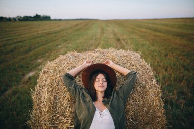 Şapkalı güzel, şık bir kadın yaz akşamı samanlığında dinleniyor. Mutlu, genç, çekici, dinlenen saman balyası. Kırsal kesimdeki atmosferik sükunet anı