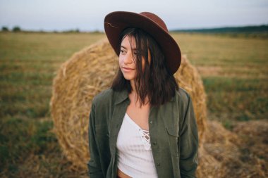 Yaz akşamları saman balyasında duran şapkalı güzel bir kadın. Genç, modaya uygun kadın portresi samanlıkta dinleniyor, yaz tatili kırsalda. Huzur