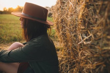 Yaz tarlasında akşam vakti samanlıkta oturan şapkalı güzel bir kadın. Genç seksi moda kadın saman balyasında gün ışığında dinleniyor. Kırsal kesimdeki atmosferik sükunet anı