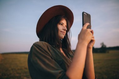 Şapkalı güzel bir kadın yaz tarlasında günbatımının fotoğrafını çekiyor. Akıllı telefon tutan genç ve çekici bir kadının portresi ve sıcak gün ışığında akşam görüntüsü. Atmosferik an