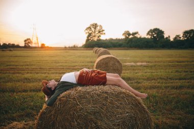 Gün batımında samanlıkta uzanmış şapkalı, kaygısız güzel bir kadın yaz tarlasında akşamın tadını çıkarıyor. Kırsalda saman balyasında rahatlayan genç ve mutlu bir dişi. Atmosferik sükunet anı