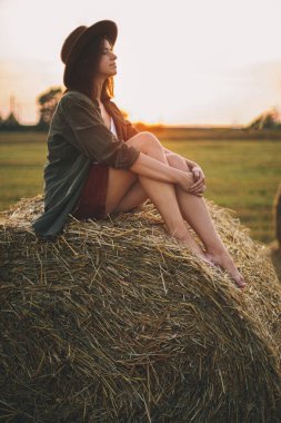 Güzel, mutlu bir kadın yaz samanlığında gün batımında samanlıkta oturuyor. Genç, mutlu bir kadın kırsalda saman balyasında dinleniyor, günışığının tadını çıkarıyor. Atmosferik sükunet anı