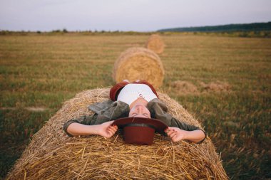 Şapkalı güzel, kaygısız kadın samanlıkta uzanıp yaz tarlasında akşamın tadını çıkarıyor. Kırsalda saman balyasında rahatlayan genç ve mutlu bir dişi. Atmosferik sükunet anı