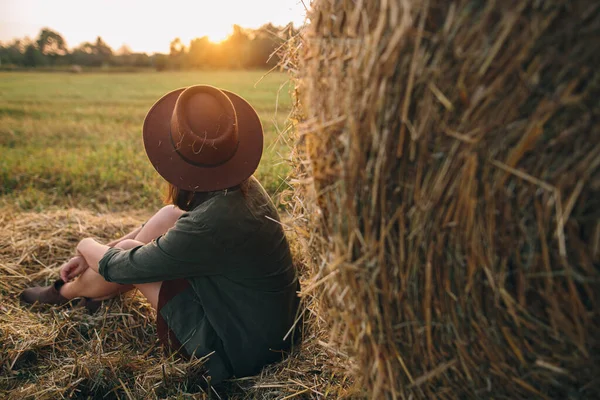 帽子のスタイリッシュな女性は 夏のフィールドで干し草に座って 日没の光を見て 田舎の静かな瞬間 暖かい日差しの中で干し草の俵で夜を楽しむ若い女性 — ストック写真
