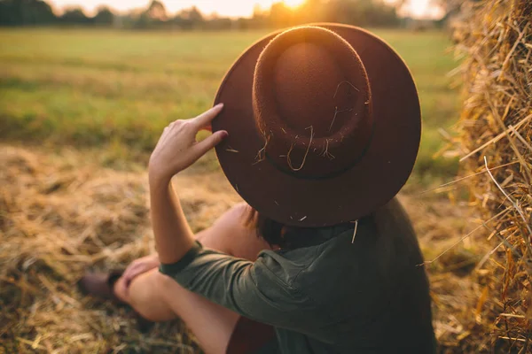日没の光を見てわらで帽子のスタイリッシュな女性は 夏のフィールドで干し草に座っています 田舎の静かな瞬間 暖かい日差しの中で干し草の俵で夜を楽しむ若い女性 — ストック写真