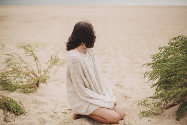 Güzel bir kadın kumlu sahilde yeşil çimlerin üzerinde oturuyor ve sakin sakin denize bakıyor. Bej örgü süveter giymiş, rüzgarlı soğuk kumsalda dinlenen zarif genç bir kadın.