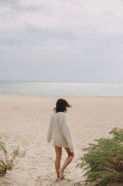 Güzel bir kadın yeşil çimlerin arkasında kumlu sahilde duruyor ve sakin sakin denize bakıyor. Bej örgü süveter giyen, rüzgarlı ve soğuk sahillerde rahatlayan zarif genç bir kadın.