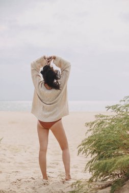 Güzel bir kadın yeşil çimlerin arkasında kumlu sahilde duruyor ve sakin sakin denize bakıyor. Bej örgü süveter giyen, rüzgarlı ve soğuk sahillerde rahatlayan zarif genç bir kadın.