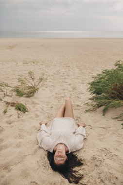 Güzel bir kadın kumlu sahilde yeşil çimlerin ve denizin arka planında uzanmış, tasasız, sakin bir an. Yukarıya bak. Örgü kazaklı şık genç bir bayan tatilin tadını çıkarıyor ve sahilde dinleniyor.