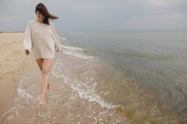 Kaygısız, örgü kazaklı güzel bir kadın soğuk deniz dalgalarında kumlu sahilde rüzgarlı saçlarıyla eğleniyor. Şık, genç, mutlu bir bayan, dinleniyor ve sahilde tatilin tadını çıkarıyor. Boşluğu kopyala