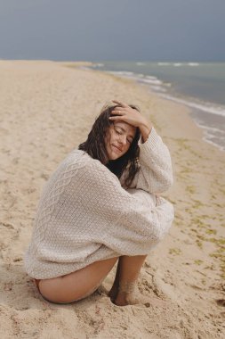 Rüzgarlı kıvırcık saçlı güzel, mutlu bir kadın denizin kumlu sahilinde oturuyor, kaygısız bir an. Sahilde rahat rahat örülü süveter giyen şık genç bir kadın. Yaz tatili