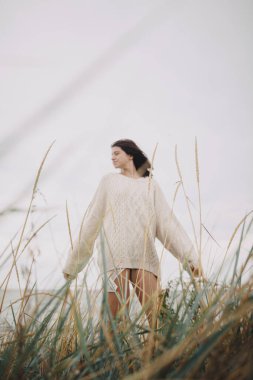 Arka planda yabani otlar örülmüş süveterli ve rüzgarlı saçlı güzel, şık bir kadının bulanık silueti. Rüzgarlı sahilde özgün kaygısız bir an. Sükunet. Dikey