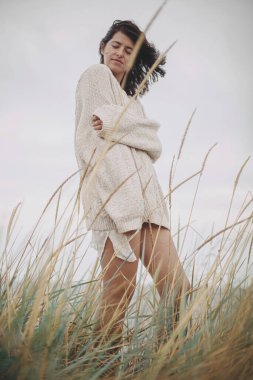 Örgü kazaklı ve rüzgarlı saçlı güzel bir kadın denizdeki yabani otların arasında poz veriyor. Otantik kaygısız an. Rüzgarlı sahilde duran şık genç bir bayan. Sükunet. Dikey