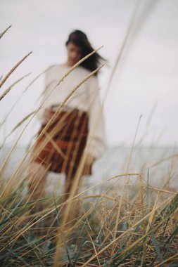 Arka planda yabani otlar örülmüş süveter giymiş güzel, şık bir kadının bulanık silueti. Rüzgarlı sahilde özgün kaygısız bir an. Sükunet. Dikey