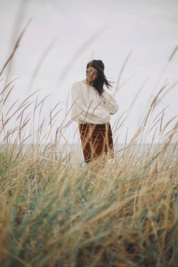 Örgü kazaklı ve rüzgarlı saçlı güzel bir kadın denizdeki yabani otların arasında poz veriyor. Otantik kaygısız an. Rüzgarlı sahilde duran şık genç bir bayan. Sükunet. Dikey