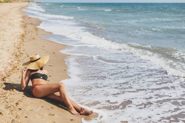 Şapkalı güzel bir kadın kumlu sahilde deniz dalgalarında oturuyor, güneşleniyor ve yaz tatilinin tadını çıkarıyor. Mesaj için yer var. Mayo ve hasır şapkalı genç bir kadın vahşi sahilde dinleniyor.
