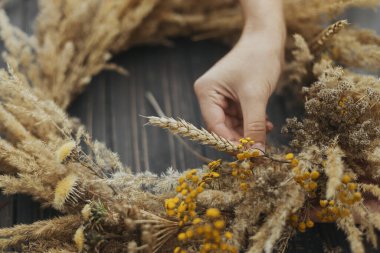 Kuru otlarla, kır çiçekleriyle ve kırsal ahşap masa üzerinde buğdayla süslü sonbahar kırsal çelenkleri yapan eller. Sonbahar tatili atölyesi. Çiçekçi koyu ağaçtan çelenk yapıyor.
