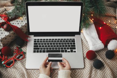 Bilgisayarın arka planında kredi kartı olan elleri, boş ekranı, şık Noel süsleri ve Noel Baba şapkası olan rahat yatağı olan elleri. Noel satışları ve online alışveriş. Metin için boşluk