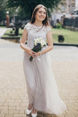 Güneşli bir sokakta kilisenin arka planında şık ve kaliteli bir elbiseyle yürüyen güzel mutlu gelin. Düğün buketi gül ve lavanta ile yürüyen duygusal gülümseyen gelin. Provence düğünü