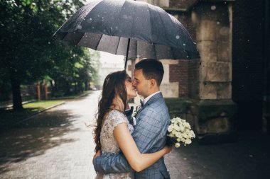 Yağmurda eski kilisenin arka planında şemsiyenin altında öpüşen buket ve damatla şık bir gelin. Yağmurlu sokakta siyah şemsiye altında kucaklaşan güzel bir çift. Romantik an
