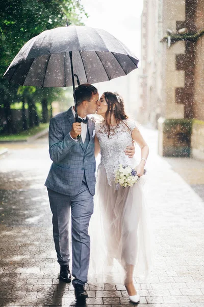 美しい結婚式のカップル一緒に傘の下で日当たりの良い雨の通りで抱擁 スタイリッシュな花嫁と新郎は雨の中で古い教会の背景に歩いてキスをします プロバンス結婚式 — ストック写真