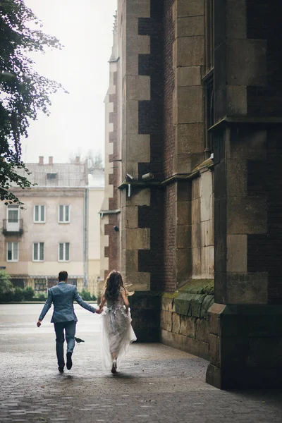 雨の通りで古い教会の背景に実行されているスタイリッシュな花嫁と新郎 プロバンス結婚式 ヨーロッパの街で雨の中で楽しんでいる美しい感情的な結婚式のカップル — ストック写真