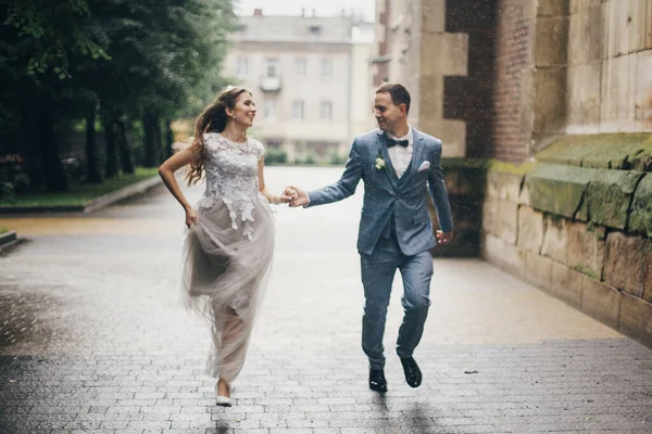 ヨーロッパの街で雨の中で笑顔の美しい感情的な結婚式のカップルのぼやけたイメージ プロバンス結婚式 雨の通りで古い教会を背景に走るスタイリッシュな幸せな花嫁と新郎 — ストック写真