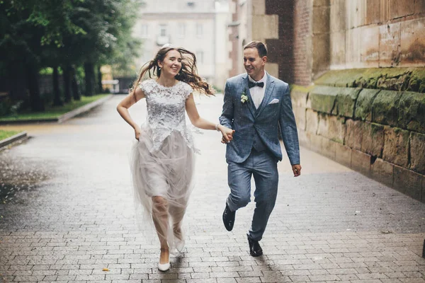 雨の通りで古い教会の背景に実行されているスタイリッシュな幸せな花嫁と新郎 プロバンス結婚式 美しい感情的な結婚式のカップル笑顔とヨーロッパの都市で雨の中で楽しみを持って — ストック写真