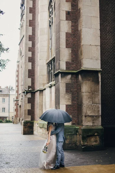 雨の中で古い教会の背景に傘の下でスタイリッシュな花嫁と新郎キス プロバンス結婚式 雨の通りで黒い傘の下で抱擁美しい結婚式のカップル ロマンチックな瞬間 — ストック写真