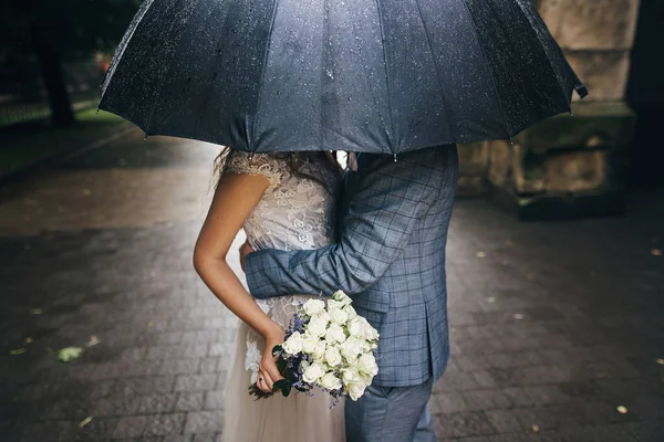 雨の中 古い教会を背景に傘の下でキスを花束と新郎とスタイリッシュな花嫁 プロバンス結婚式 美しい結婚式のカップル受容下黒傘で雨の通り — ストック写真