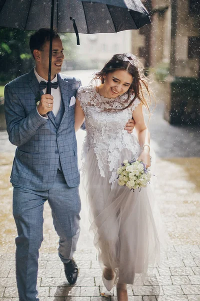 美しい幸せな結婚式のカップル一緒に傘の下で日当たりの良い雨の通りで抱擁 スタイリッシュな花嫁と新郎は雨の中で古い教会の背景に歩いて笑顔 プロバンス結婚式 — ストック写真