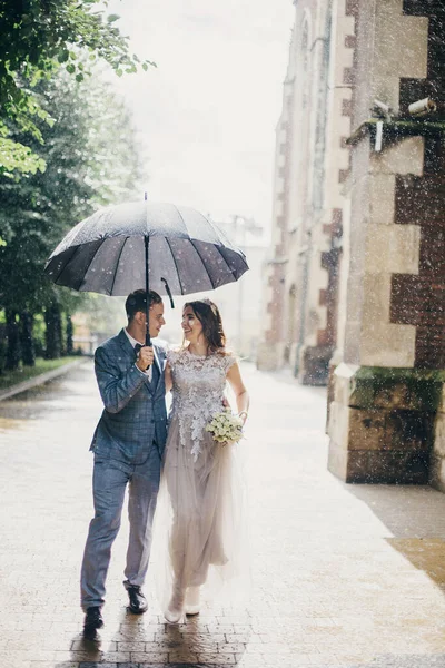 スタイリッシュな幸せな花嫁と新郎歩くと雨の中で古い教会の背景に笑みを浮かべて プロバンス結婚式 美しい結婚式のカップル一緒に傘の下で日当たりの良い雨の通り — ストック写真