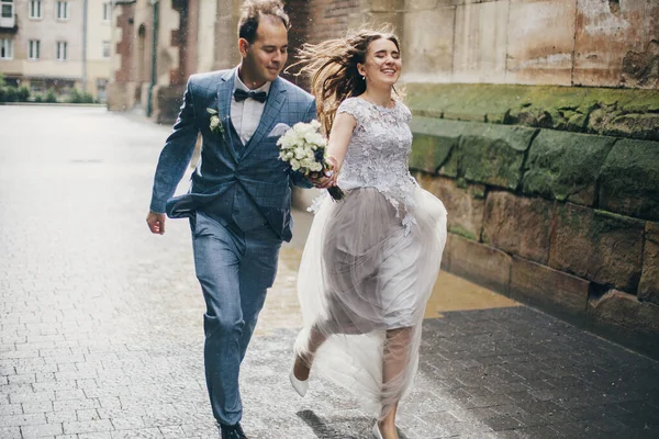 美しい感情的な結婚式のカップルは笑顔で ヨーロッパの都市で雨の中で楽しんでいます プロバンス結婚式 雨の通りで古い教会を背景に走るスタイリッシュな幸せな花嫁と新郎 — ストック写真
