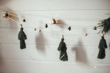 Noel süsleri, el yapımı ağaçlar ve Noel ışıkları olan beyaz ahşap duvarda çanlar. Şenlikli dekore edilmiş atmosferik İskandinav odası. Boho basit el yapımı dekorasyonlar