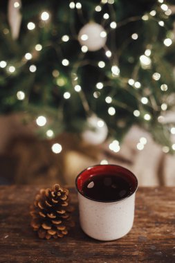 Tarz bardağı ve çam kozalağı kırsal ahşap üzerinde akşam odasındaki ışıklarda Noel ağacının arka planında. Mesaj için yer var. Dekoratif odada çay ile seramik bardak. İyi tatiller. Atmosfer zamanı
