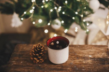 Tarz bardağı ve çam kozalağı kırsal ahşap üzerinde akşam odasındaki ışıklarda Noel ağacının arka planında. Mesaj için yer var. Dekoratif odada çay ile seramik bardak. İyi tatiller. Atmosfer zamanı