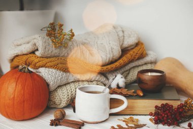 Merhaba sonbahar, rahat ve yavaş yaşam. Balkabağı, bir fincan çay, rahat süveterler, sonbahar yaprakları, yanan mum ve klasik kitap altın ışıklarıyla dolu, beyaz ahşap arka planda. Mutlu Şükran Günleri