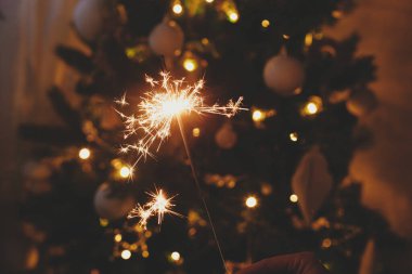 Yeni yılınız kutlu olsun! Noel ağacı ışıklarının arka planında yanan maytap. Havai fişek bangının şenlikli akşam odasında parlaması. Mesaj için yer var. Atmosferik an. Mutlu Noeller ve mutlu tatiller.