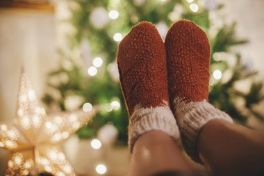 Noel ağacının arka planında, rahat yün çoraplar içindeki kadın ayakları ve şenlikli akşam odasındaki ışıklarda yıldız. Evdeki sıcacık kış anları. Kadın bacaklarında şık, sıcak çoraplar.